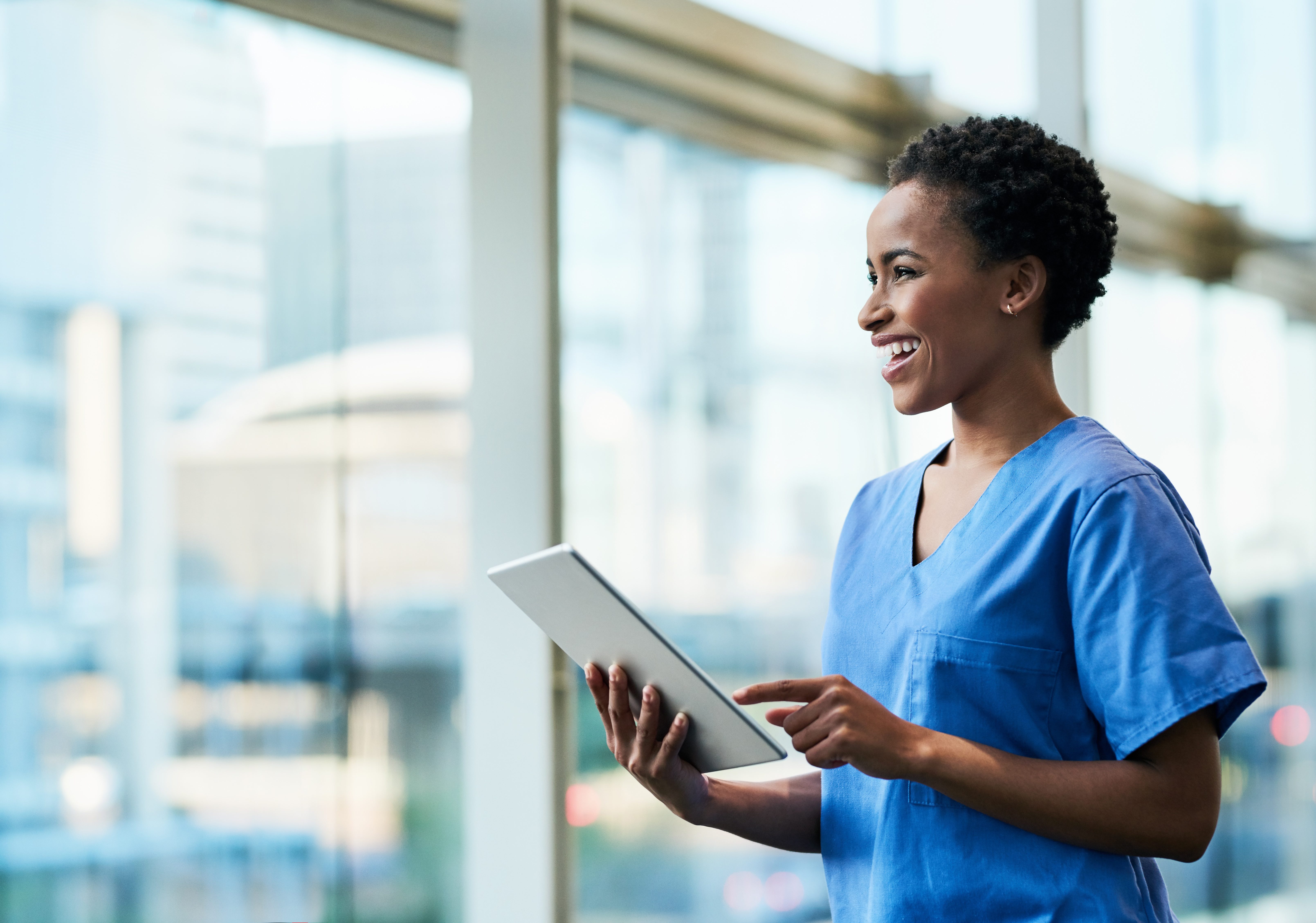 medical provider in blue scrubs holding a tablet and smiling in front of a window