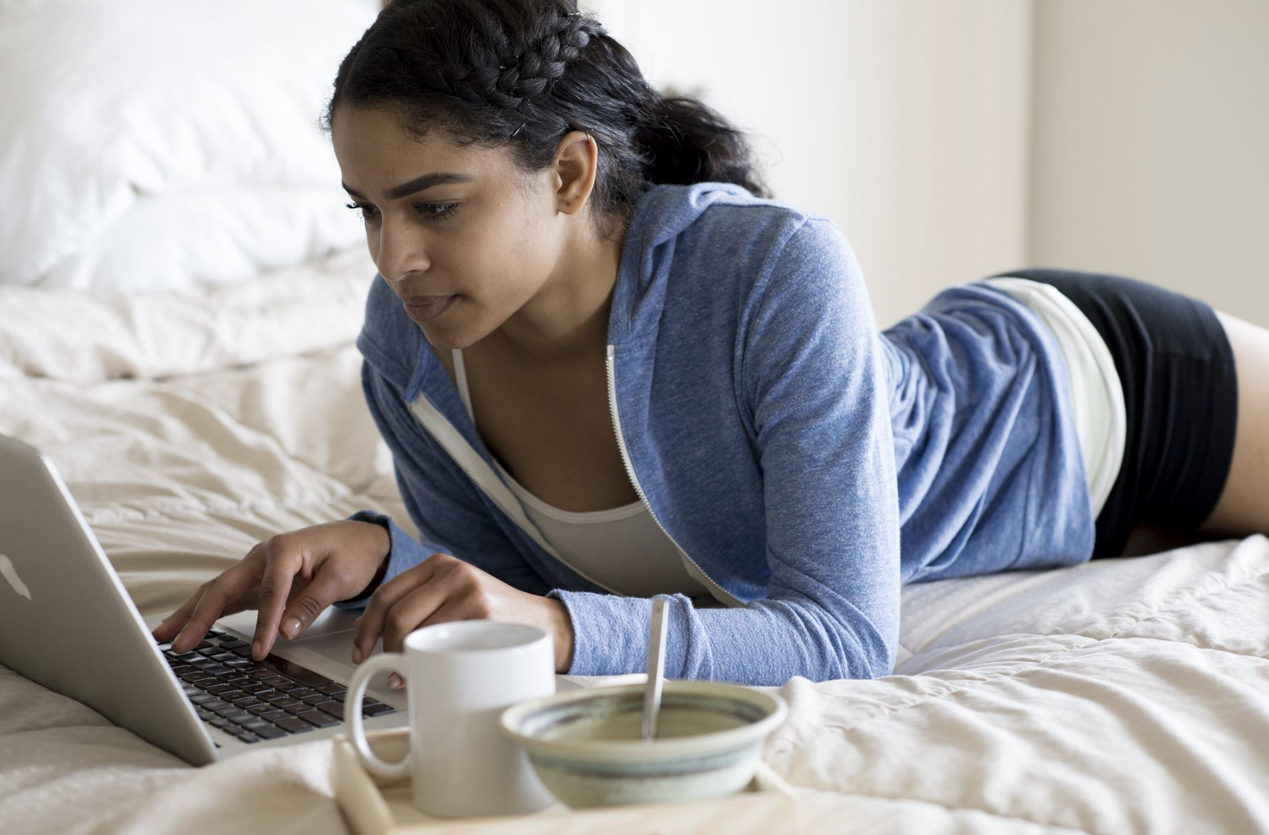 woman with dark hair and a blue sweatshirt using her laptop on her bed