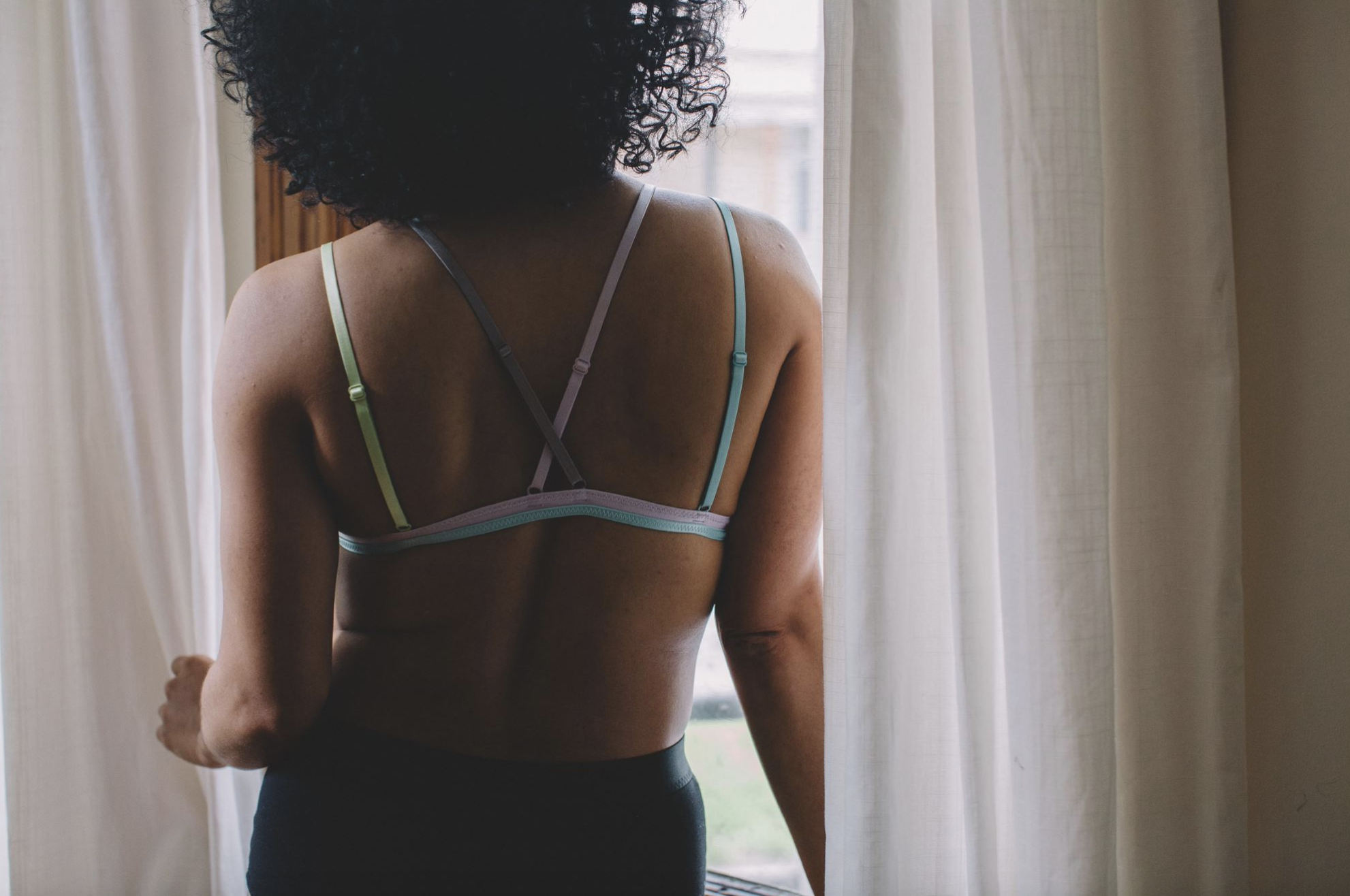 black woman in a bra standing by a window and looking out