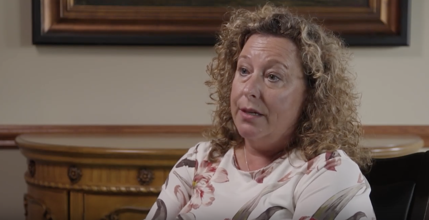 white woman with blonde curly hair and a floral white top being interviewed