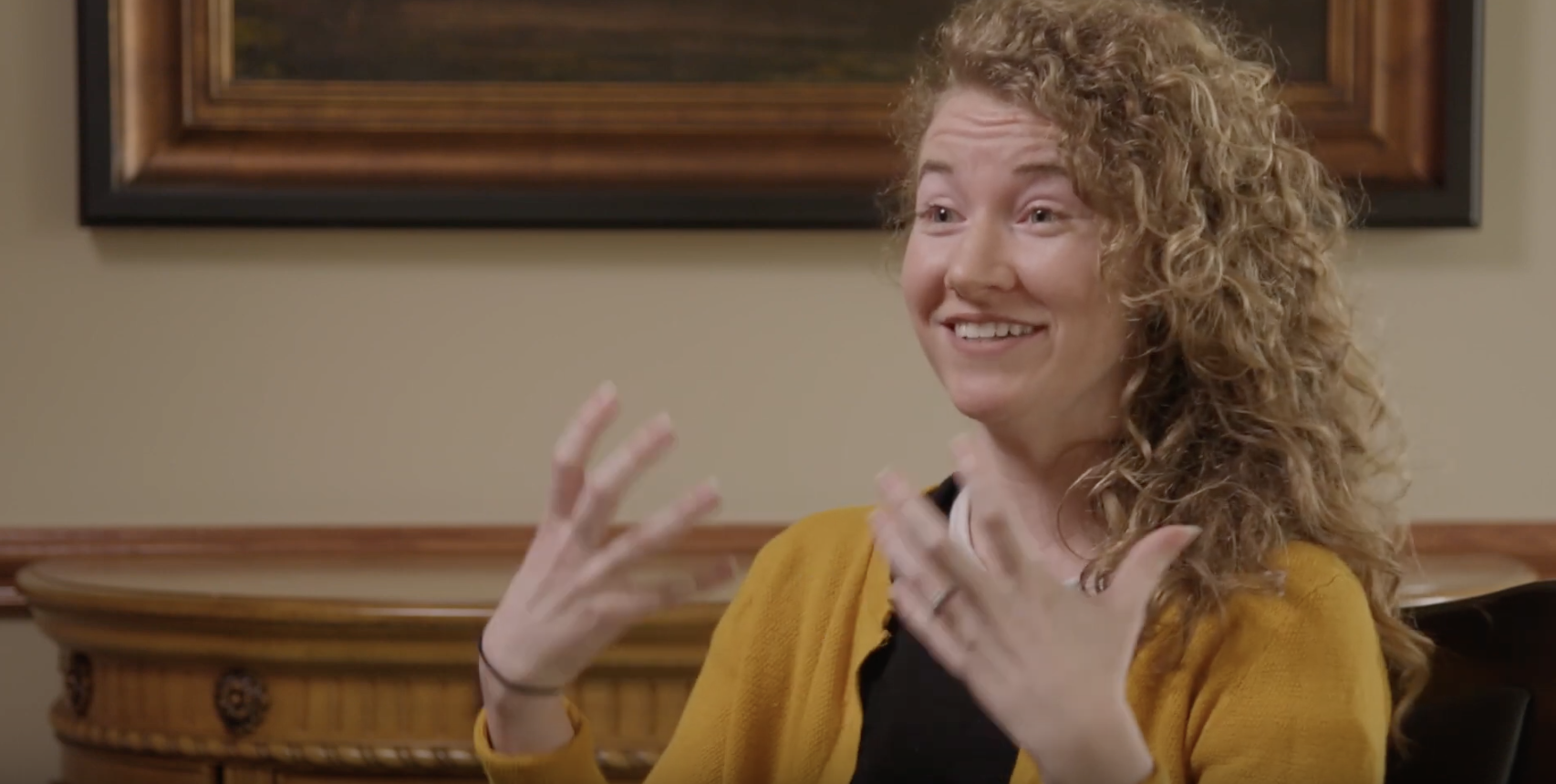 white woman with blonde curly hair in a black shirt and a yellow cardigan being interviewed