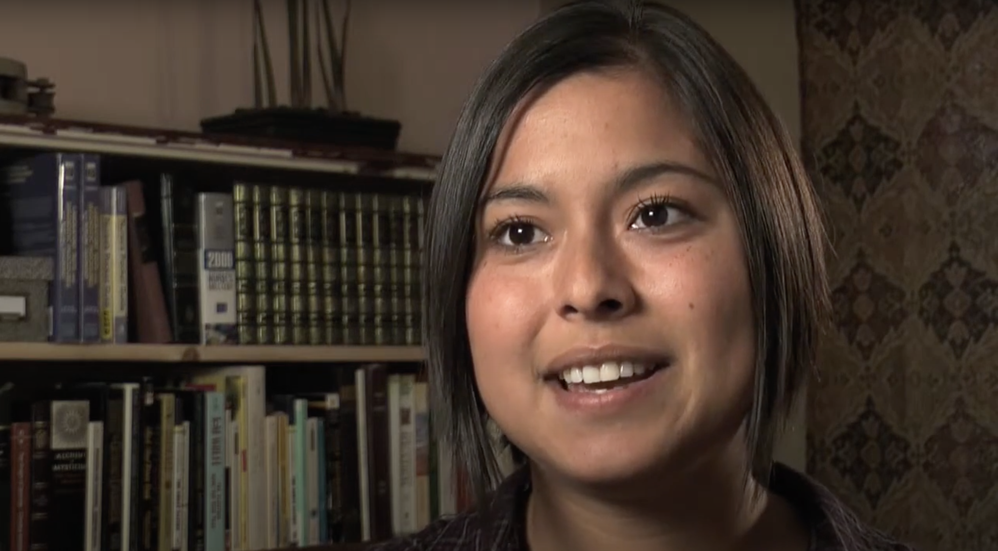 BIPOC woman with dark hair talking about her birth control with books on a bookshelf in the background