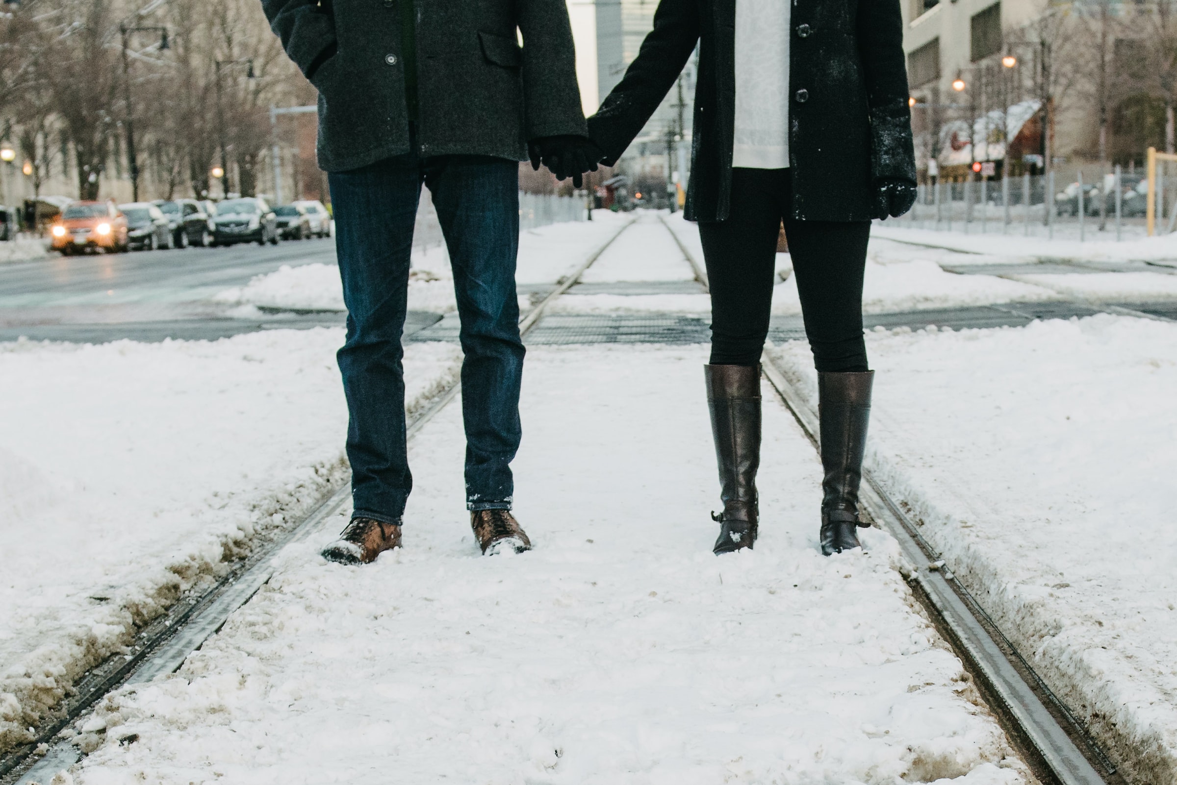 couple shown from the shoulders down holding hands while standing in a city with snow on the ground