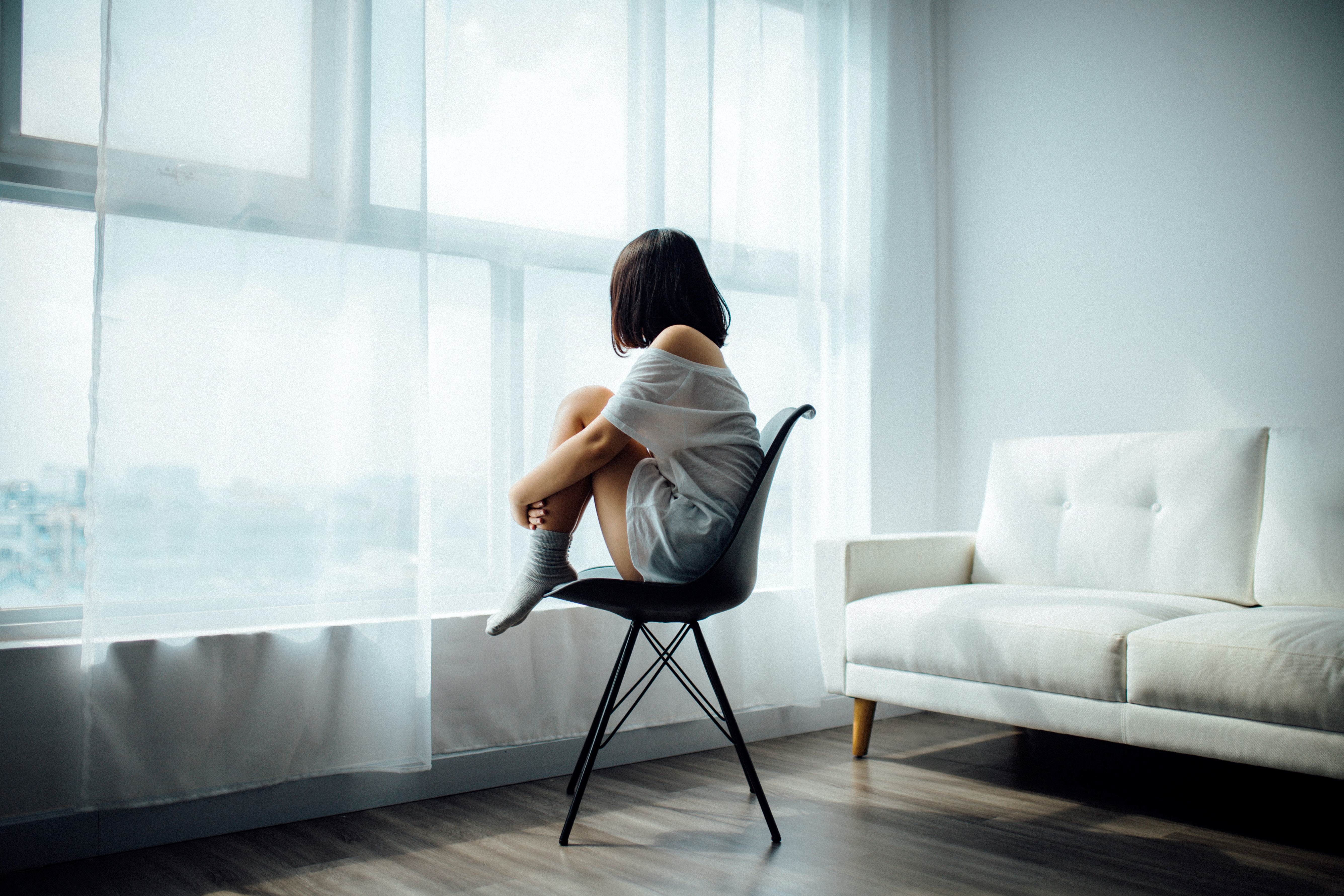 woman in a white shirt sitting in a black chair in front of a window, looking out with her knees pulled into her chest