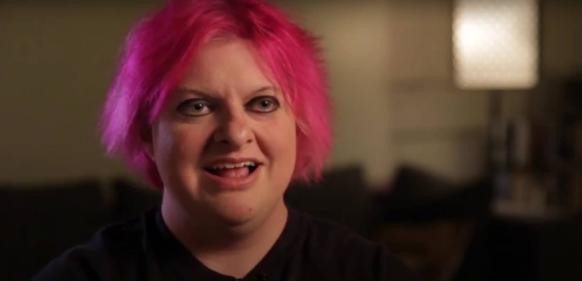 person wearing a dark shirt with pink hair talking to the camera