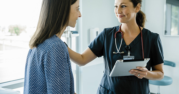 female medical provider holding a clipboard and comforting a female patient wearing an exam gown