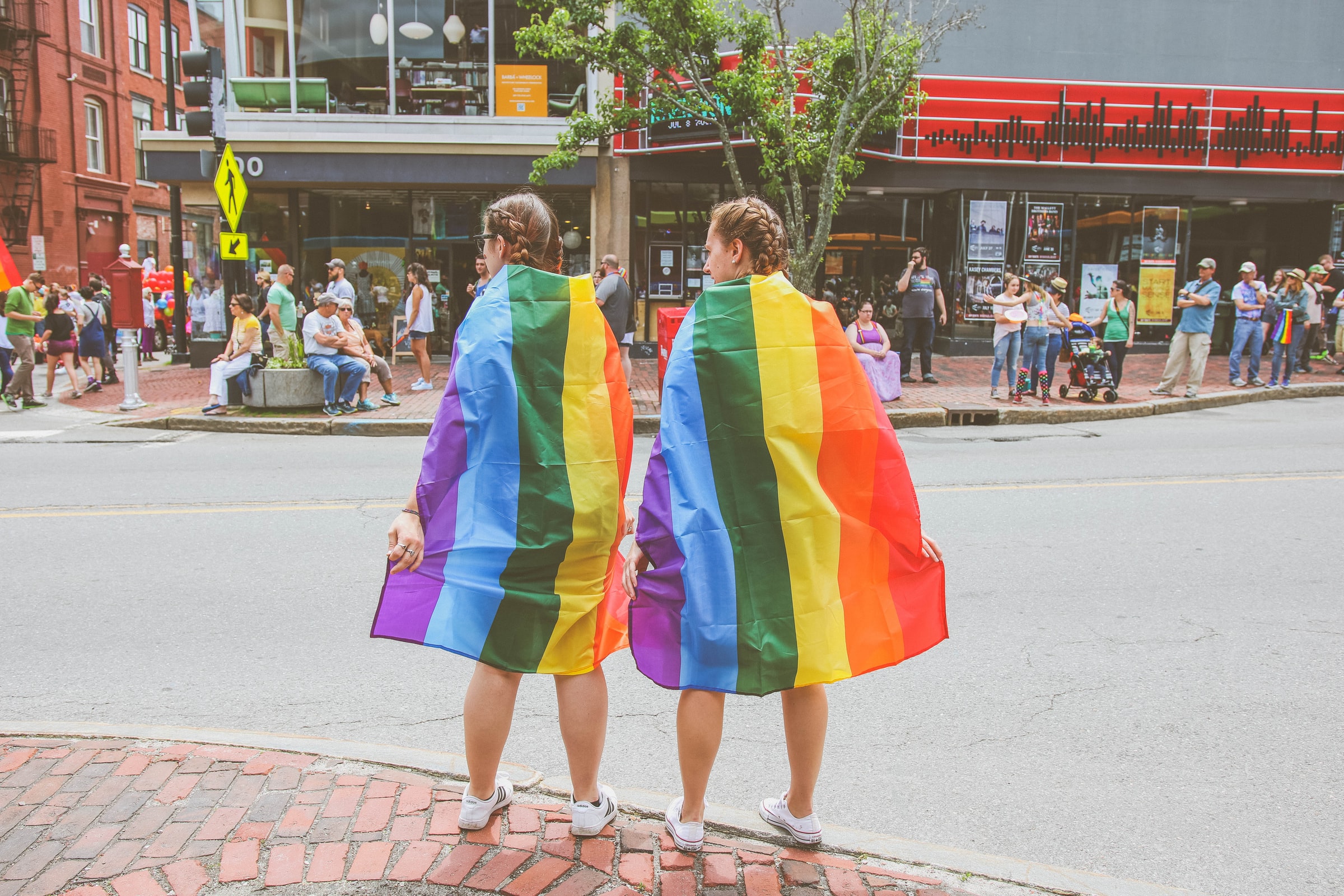 two people wearing pride flags around their shoulders waiting to cross a city street