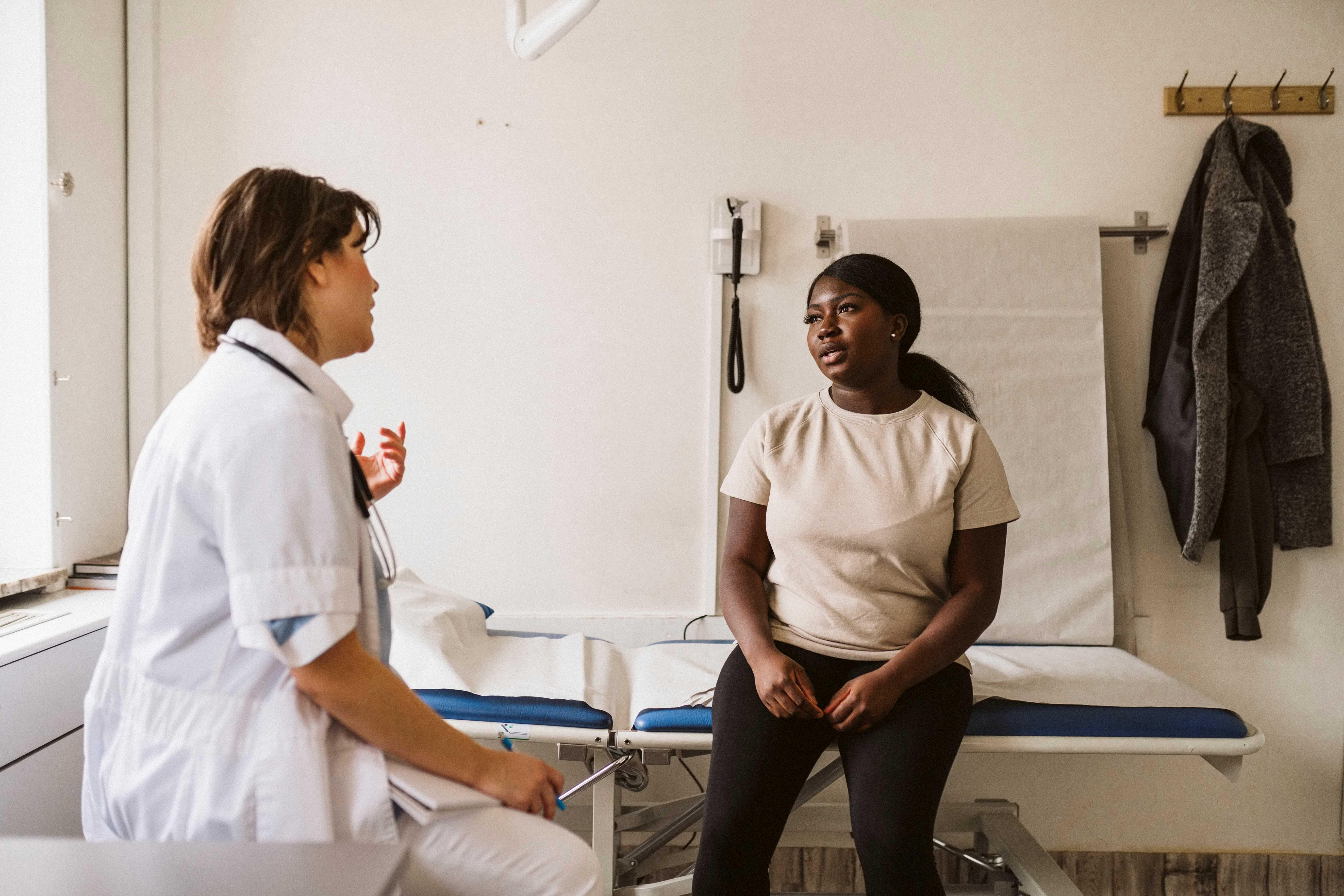 young bipoc female sitting on table in an exam room, talking to a provider in a white coat