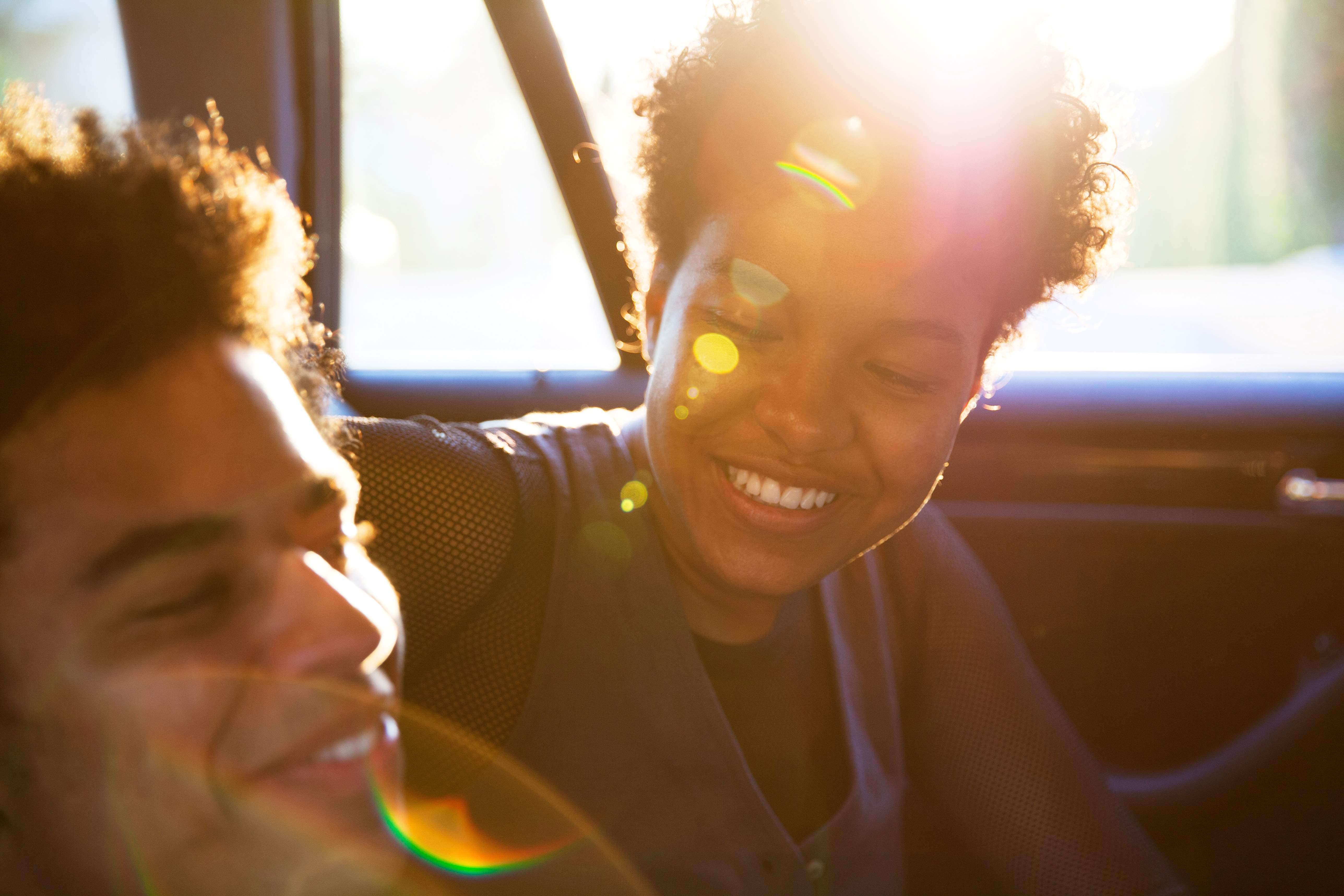 BIPOC female and male sitting in the back of a car laughing with the sun shining