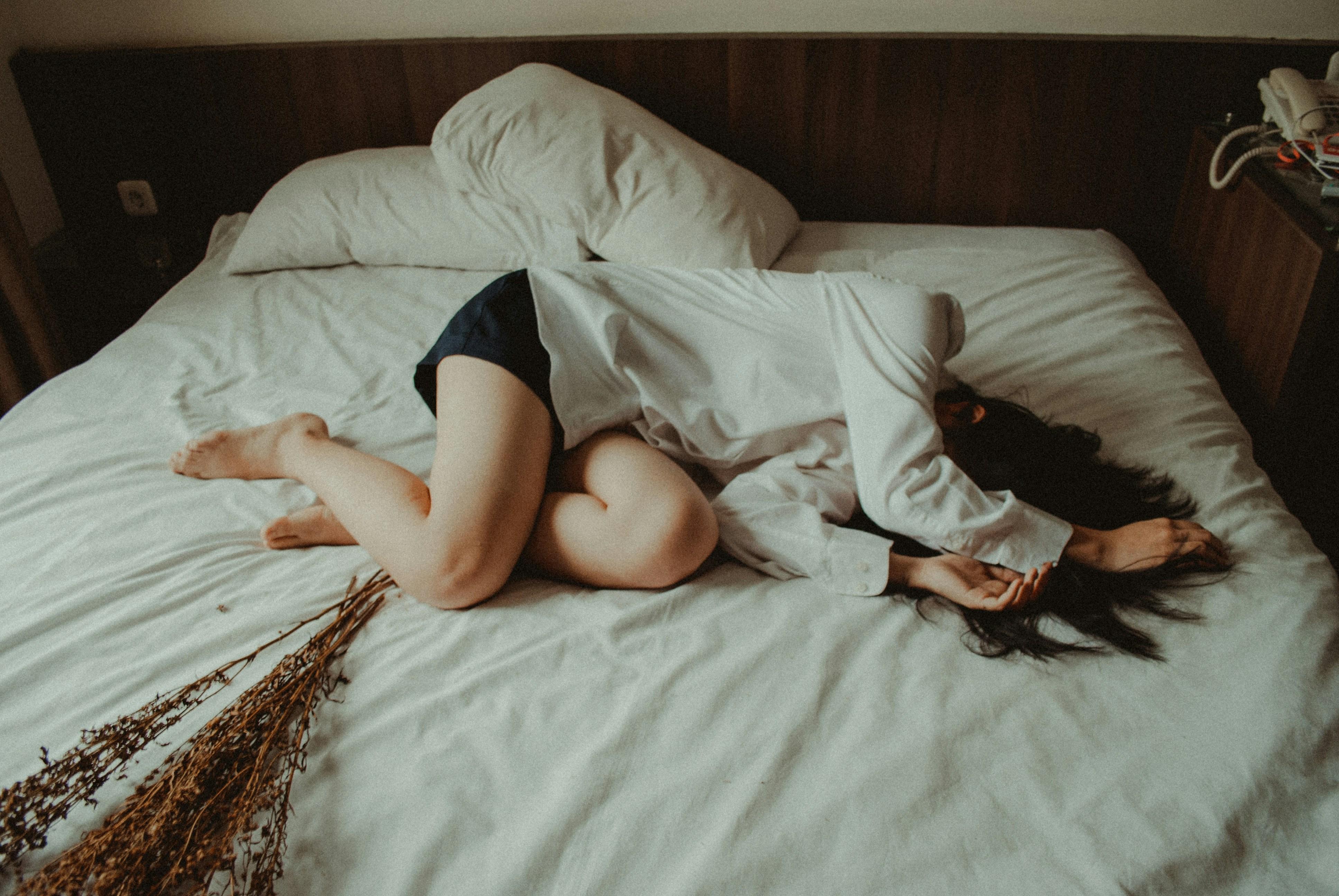 Woman lying curled up on a bed in a dimly lit room, wearing a white shirt and dark shorts, with her face turned away and dried flowers resting nearby.