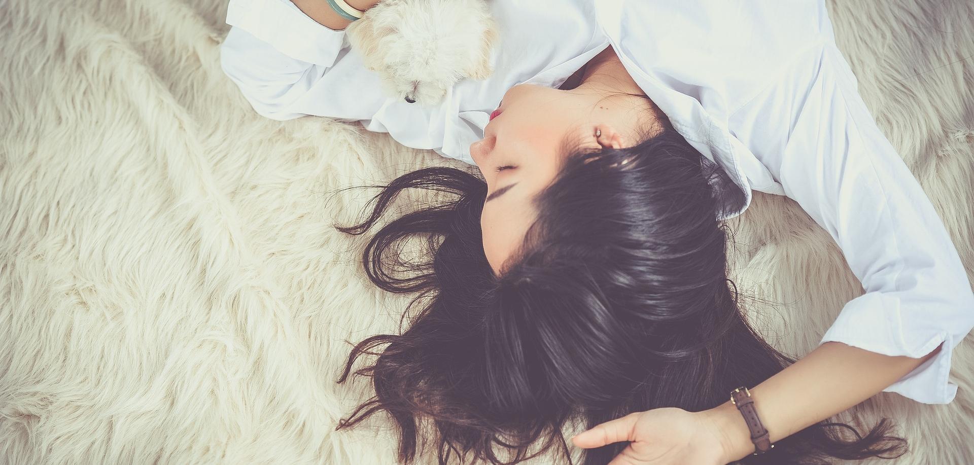 woman with long dark hair lying on bed with small white dog