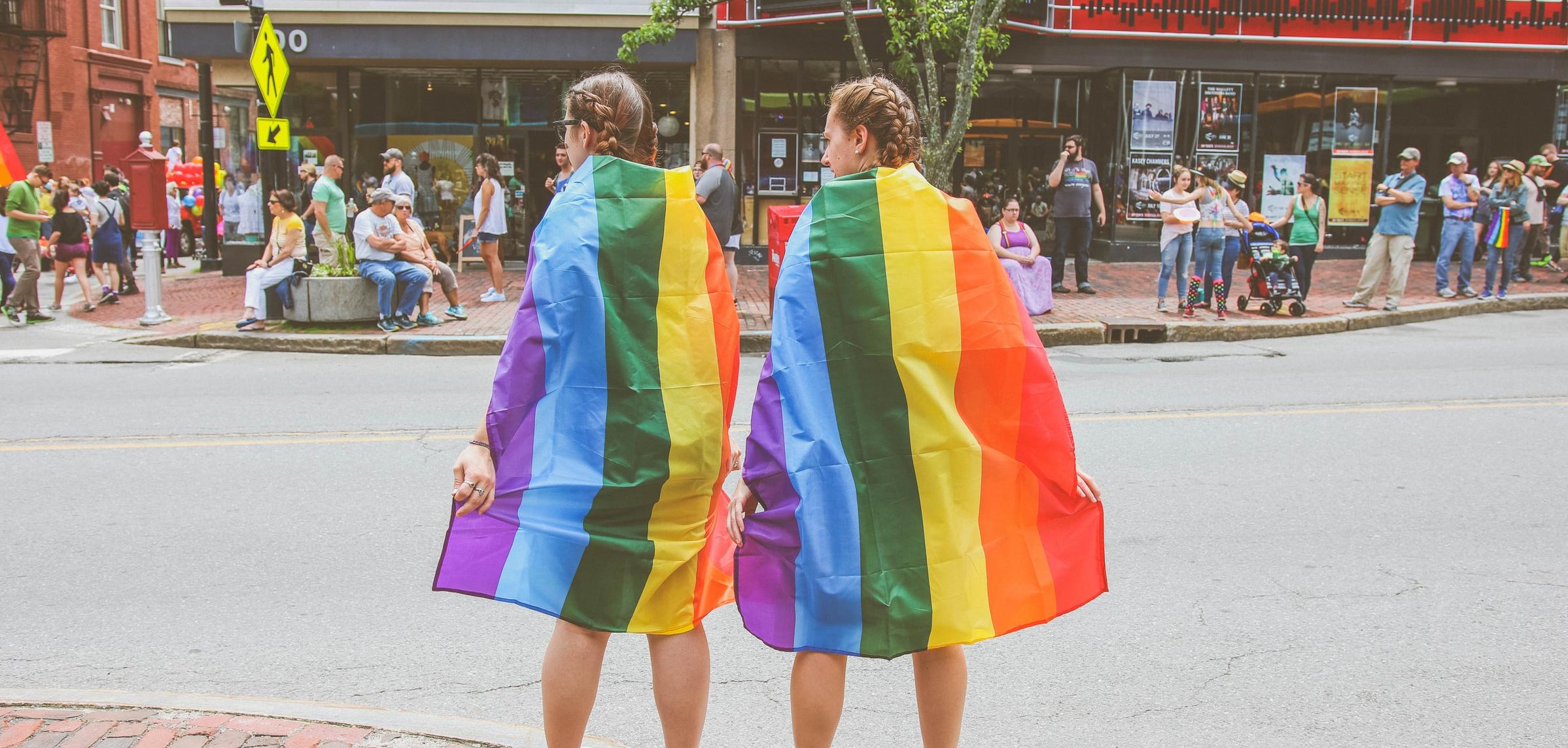 two people wearing pride flags around their shoulders waiting to cross a city street