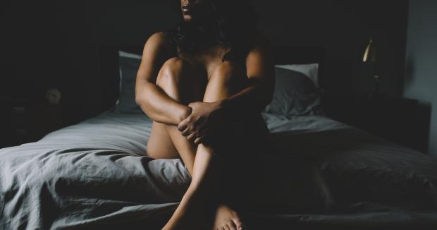 woman sitting on the edge of a bed in a dark room with her knees pulled up to her chest