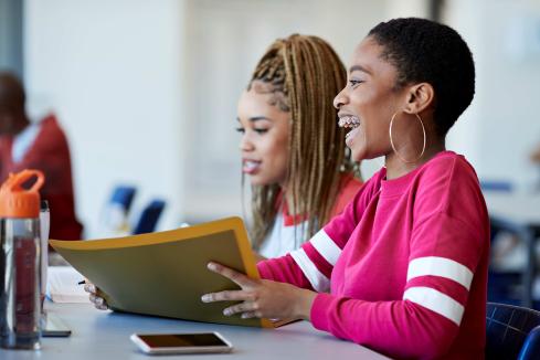 two female poc students sitting at a table near one cell phone, one is smiling and holding a folder, perhaps homework