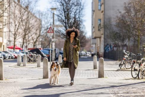 young BIPOC woman walking her dog on the street in a city, looking at her cell phone