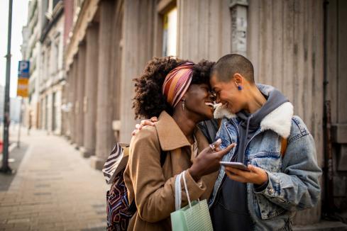 two BIPOC people embracing on the street, laughing and smiling, one holding a shopping bag and the other holding a cell phone