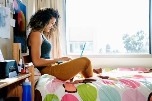 young female POC sitting on a polka dot bedspread using a laptop next to a window in dorm