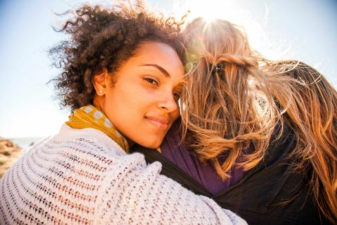 young bipoc woman facing the camera, side hugging a friend with long blonde hair who is facing away