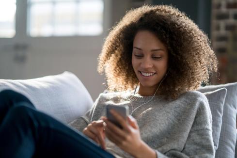 young bipoc woman in a gray sweatshirt sitting on a couch using her phone with headphones on and smiling