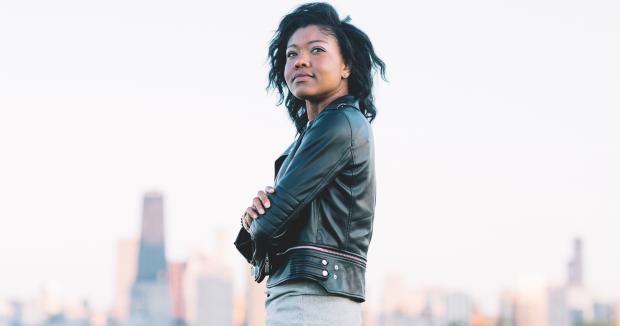 BIPOC woman in leather jacket and jeans looking off into the distance with a cityscape behind her