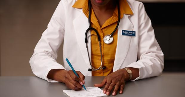 BIPOC female medical provider wearing a yellow shirt and a white coat writing on a notepad with a blue pen
