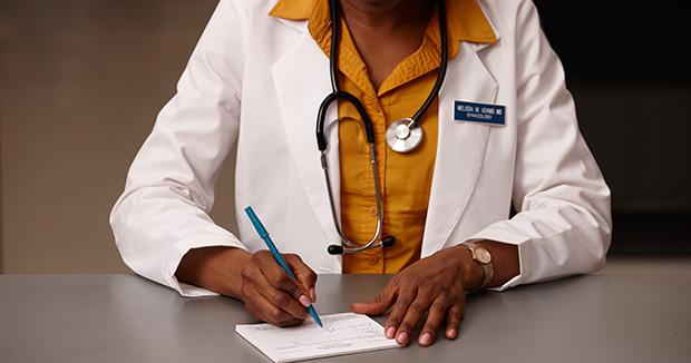 BIPOC female medical provider wearing a yellow shirt and a white coat writing on a notepad with a blue pen