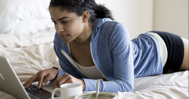 woman with dark hair and a blue sweatshirt using her laptop on her bed