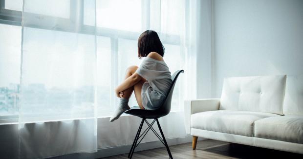 woman in a white shirt sitting in a black chair in front of a window, looking out with her knees pulled into her chest