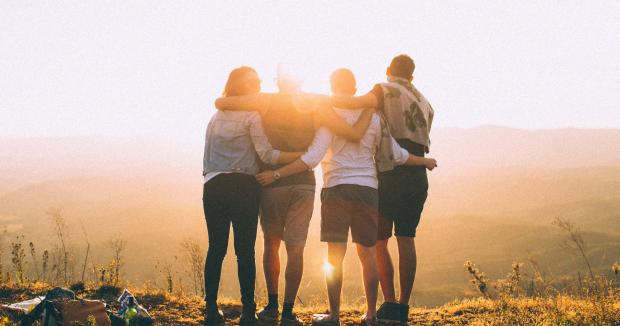 four friends with their arms around each other on a mountain at sunset