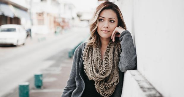 woman with brown hair standing on the street leaning against a building and looking into the distance