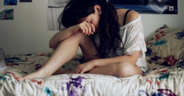 Woman with long dark hair  sitting on a bed, resting her head and her arm on her knee