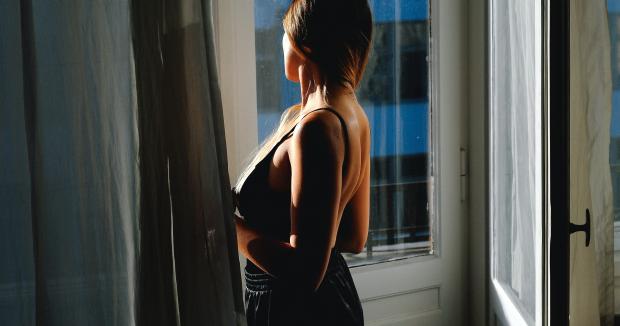 woman with long brown hair in a tanktop and jeans looking out the window