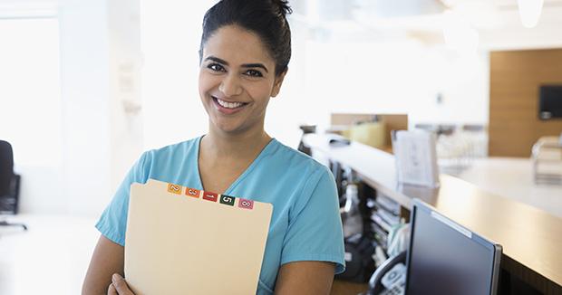 smiling female medical provider wearing scrubs holding a folder while standing in an examination room