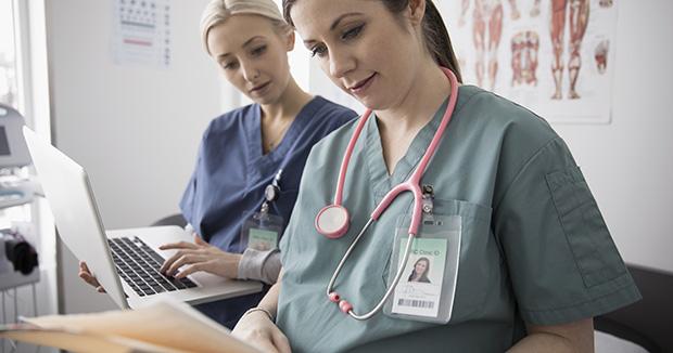 two female medical providers wearing scrubs, one holding a laptop and the other looking at a medical file