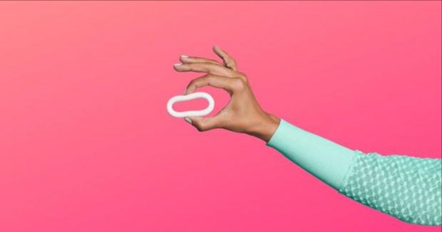 an arm holding up birth control ring with two fingers against a pink background