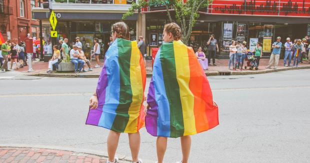 two people wearing pride flags around their shoulders waiting to cross a city street