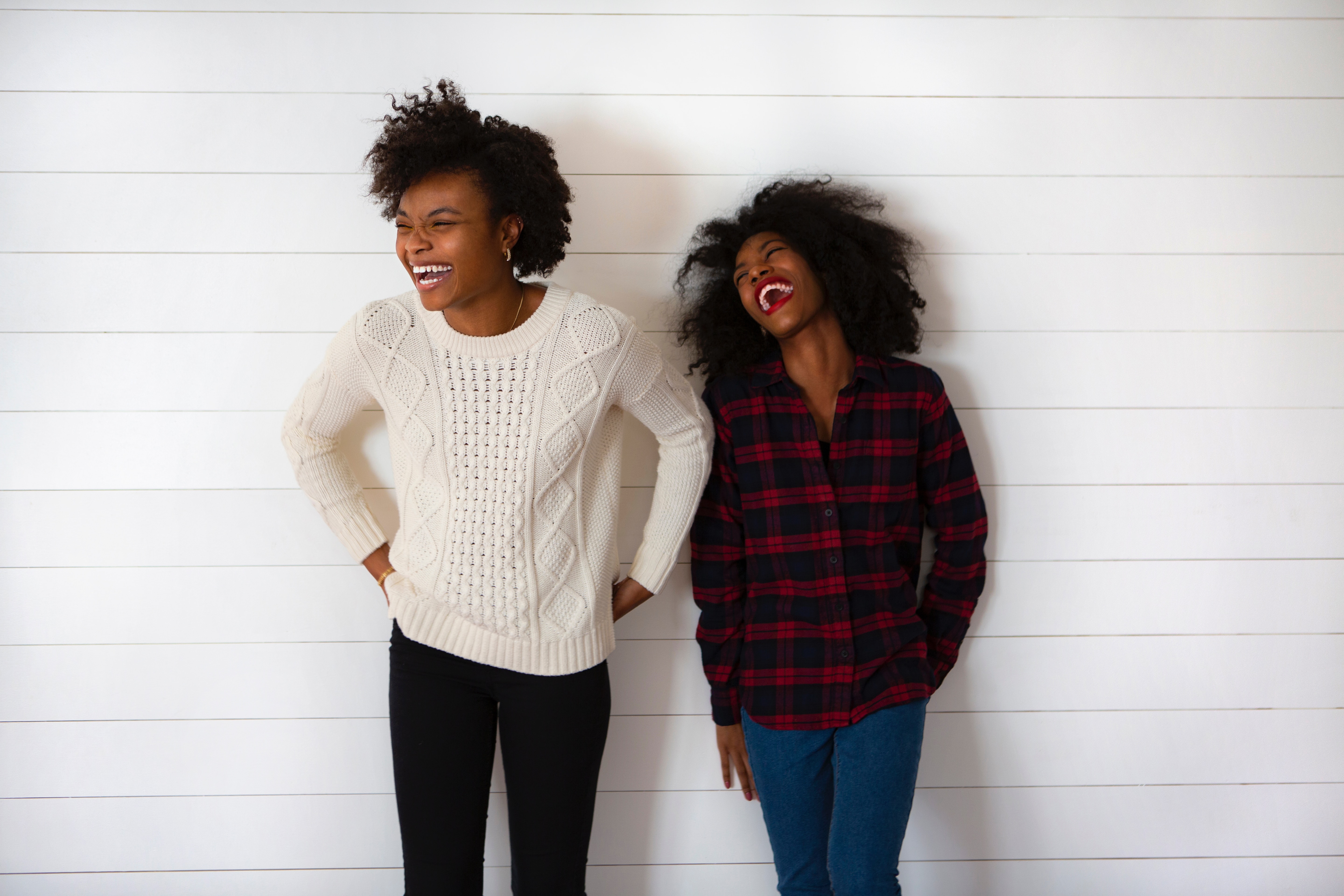 two black women standing against a white wall and laughing