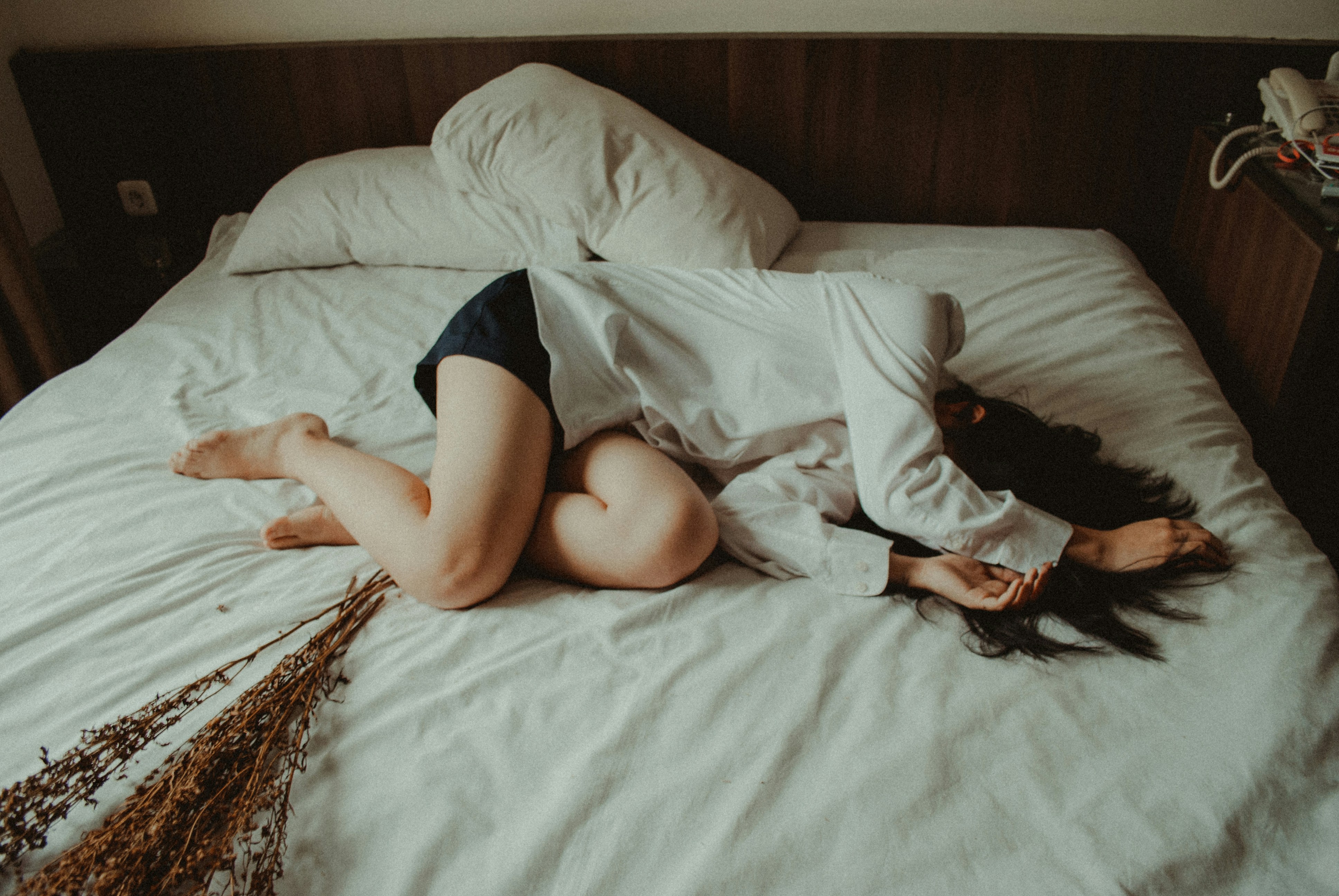 Woman lying curled up on a bed in a dimly lit room, wearing a white shirt and dark shorts, with her face turned away and dried flowers resting nearby.
